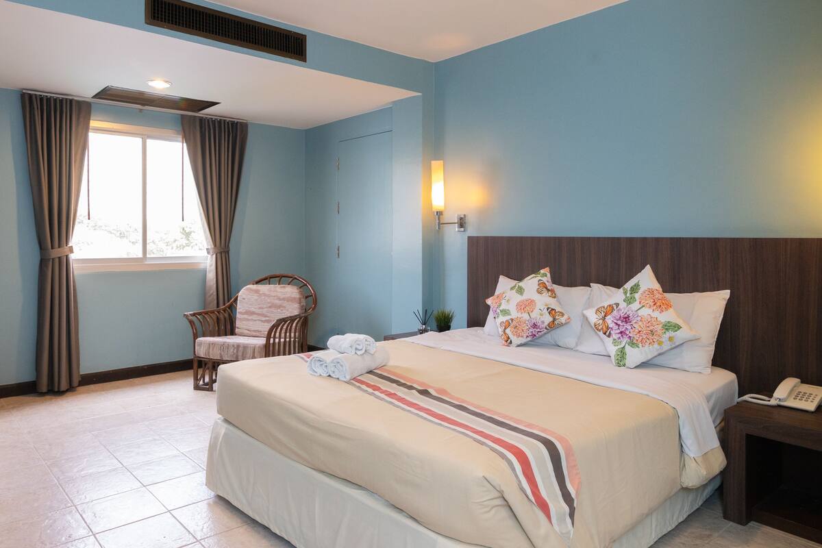 A hotel room with a large bed, a chair, a bedside table with a phone, a window with curtains, and a wall-mounted light.