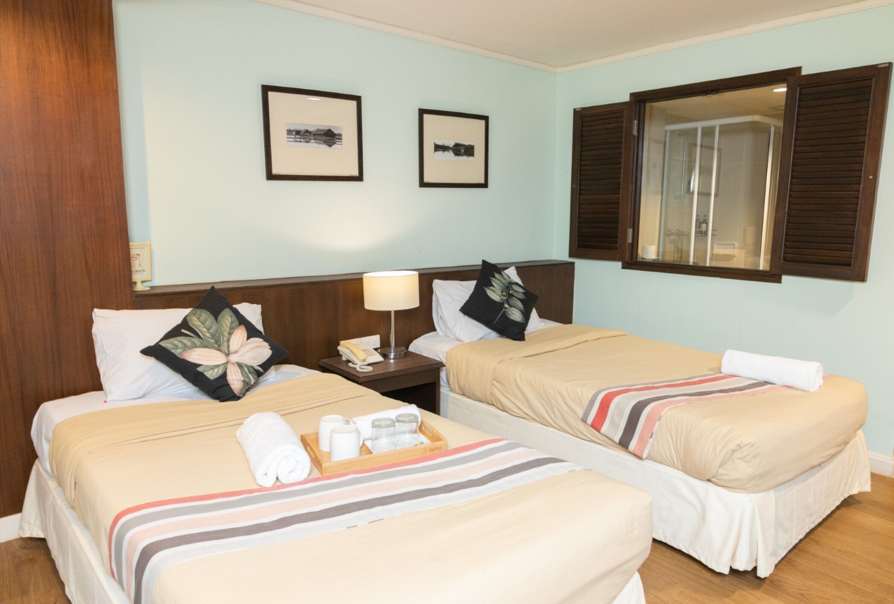 A hotel room with two beds, a wooden headboard, a nightstand with a lamp, framed pictures on the wall, and a window with shutters.