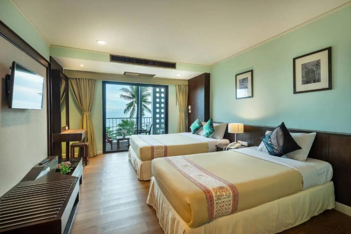 A hotel room with two beds, a TV, a desk, and a balcony with a view of palm trees.