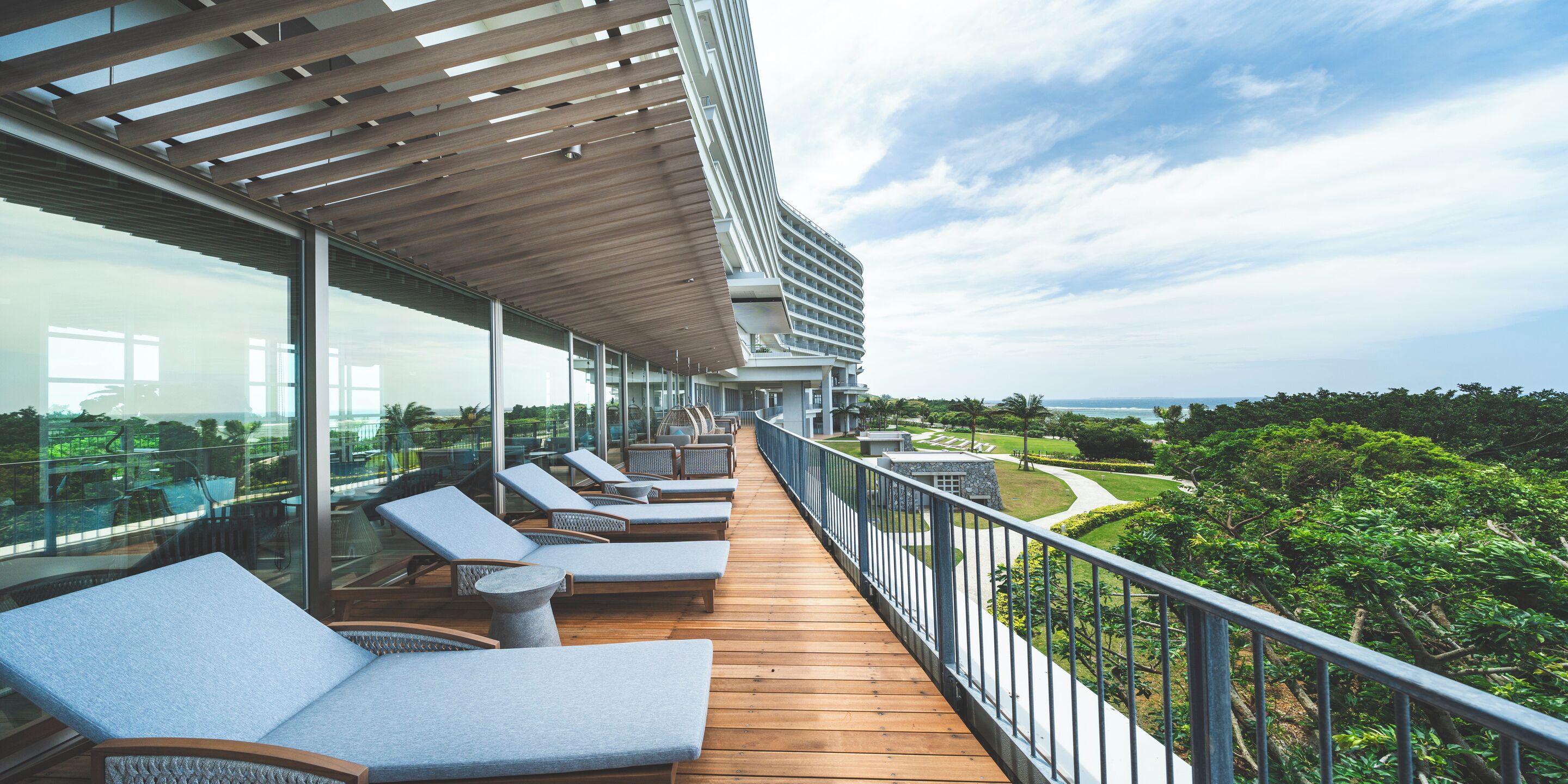 Terraza con sillones y barandilla de vidrio con vista a un jardín ajardinado y al mar.