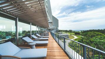Terraza con sillones y barandilla de vidrio con vista a un jardín ajardinado y al mar.