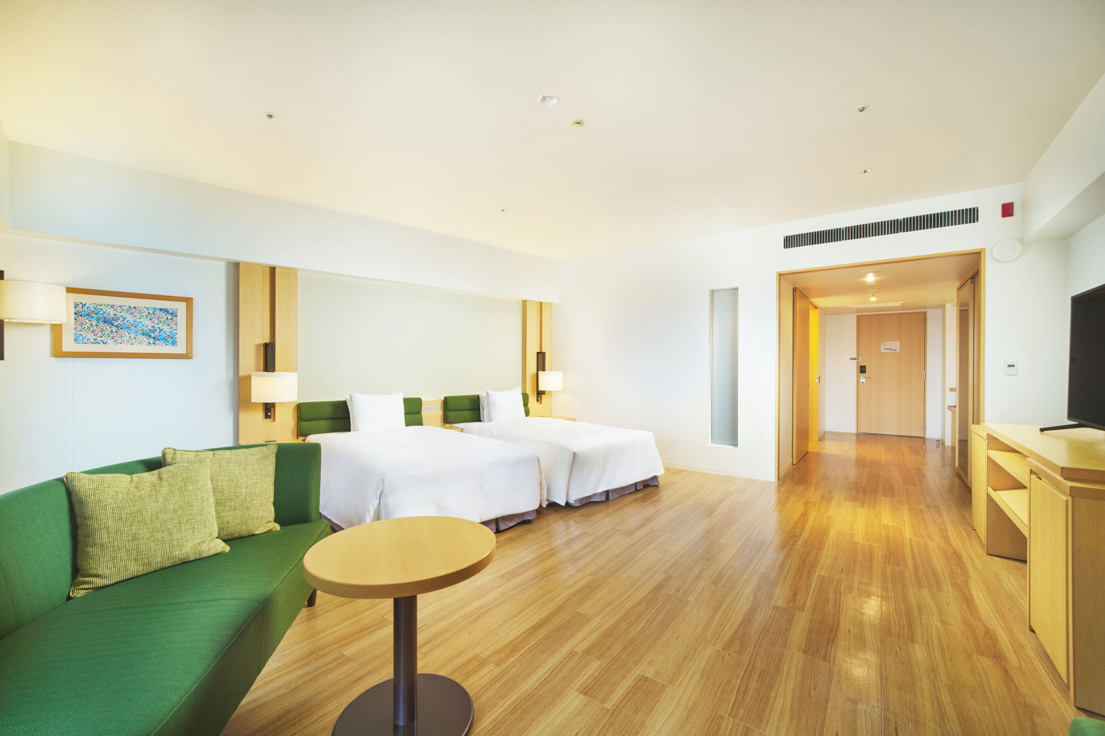 A hotel room with a green sofa, two single beds, a wooden floor, a small round table, and a TV on a wooden cabinet.
