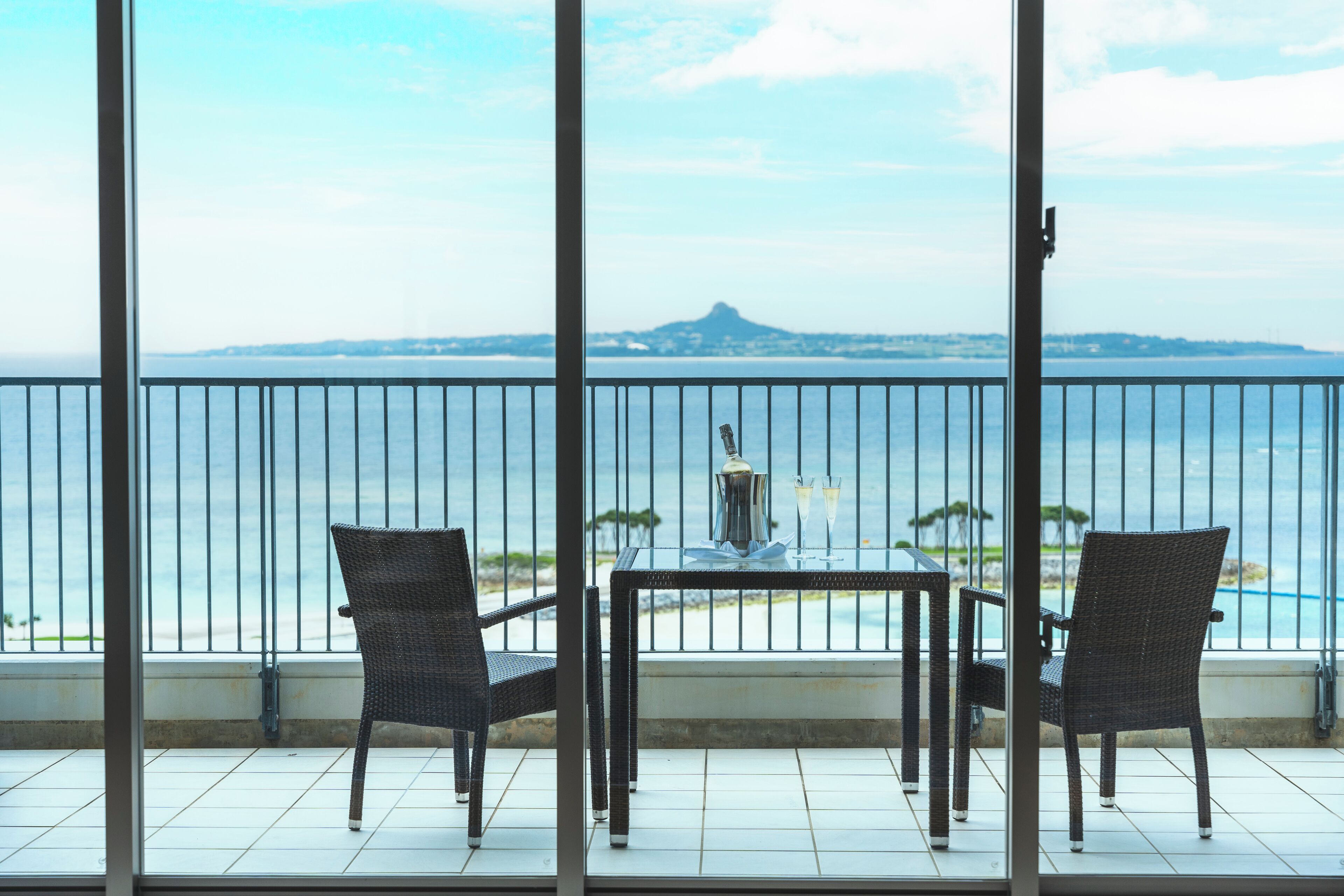 Ein Balkon mit zwei Korbstühlen und einem Tisch, der auf ein Gewässer und ein fernes Gebirge blickt.