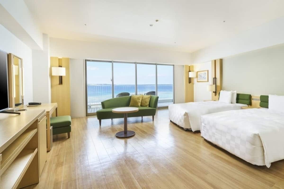 Une chambre d’hôtel avec une grande fenêtre, deux lits, un bureau et un fauteuil vert.