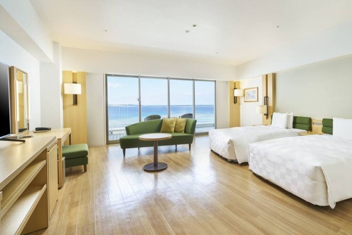 A hotel room with a large window, two beds, a desk, and a green armchair.