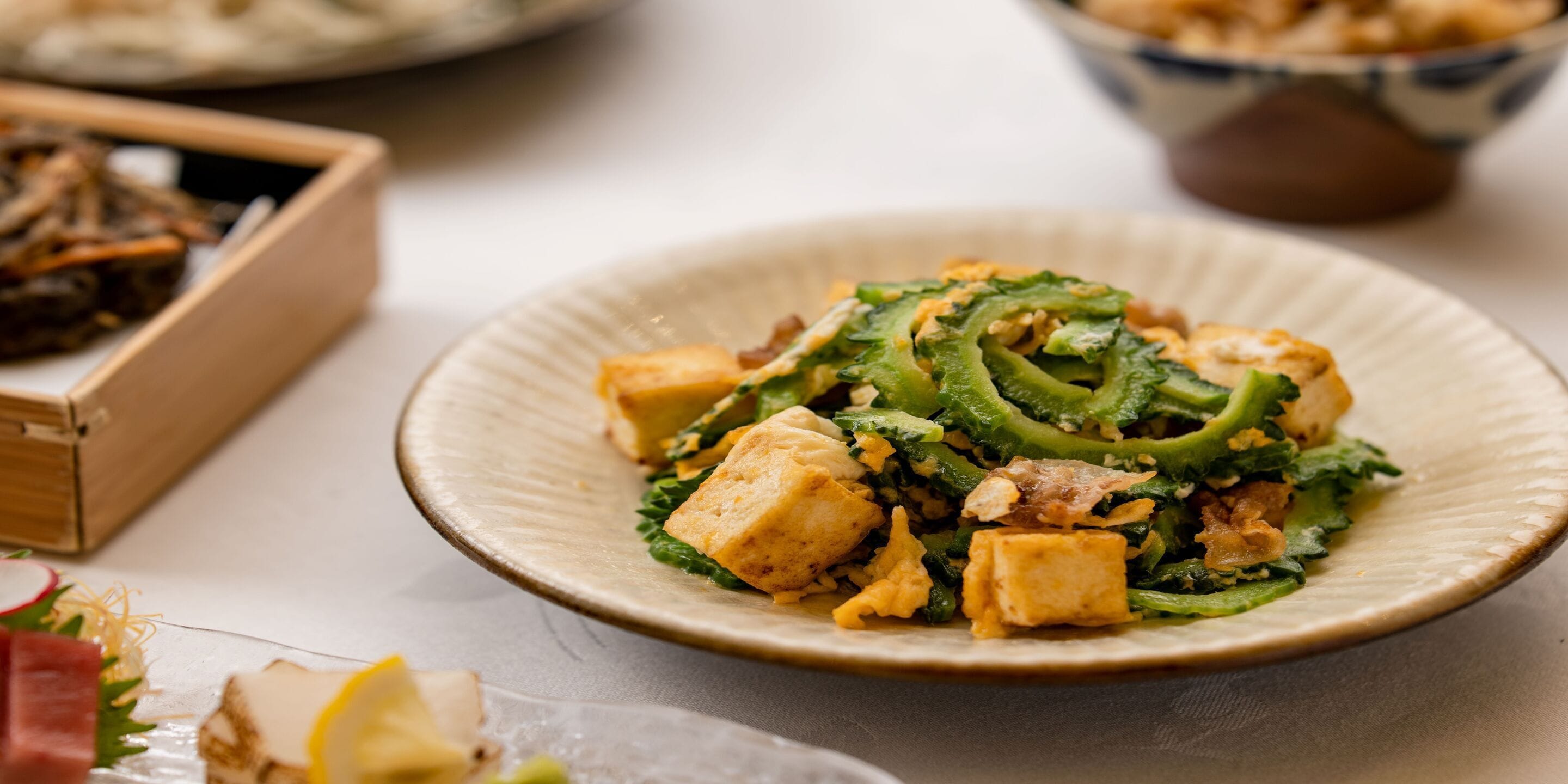 A plate of food with tofu and greens, a wooden box, and a bowl in the background.