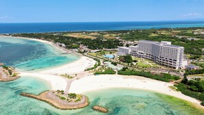 An aerial view of a coastal hotel with a beachfront, clear waters, and lush greenery.