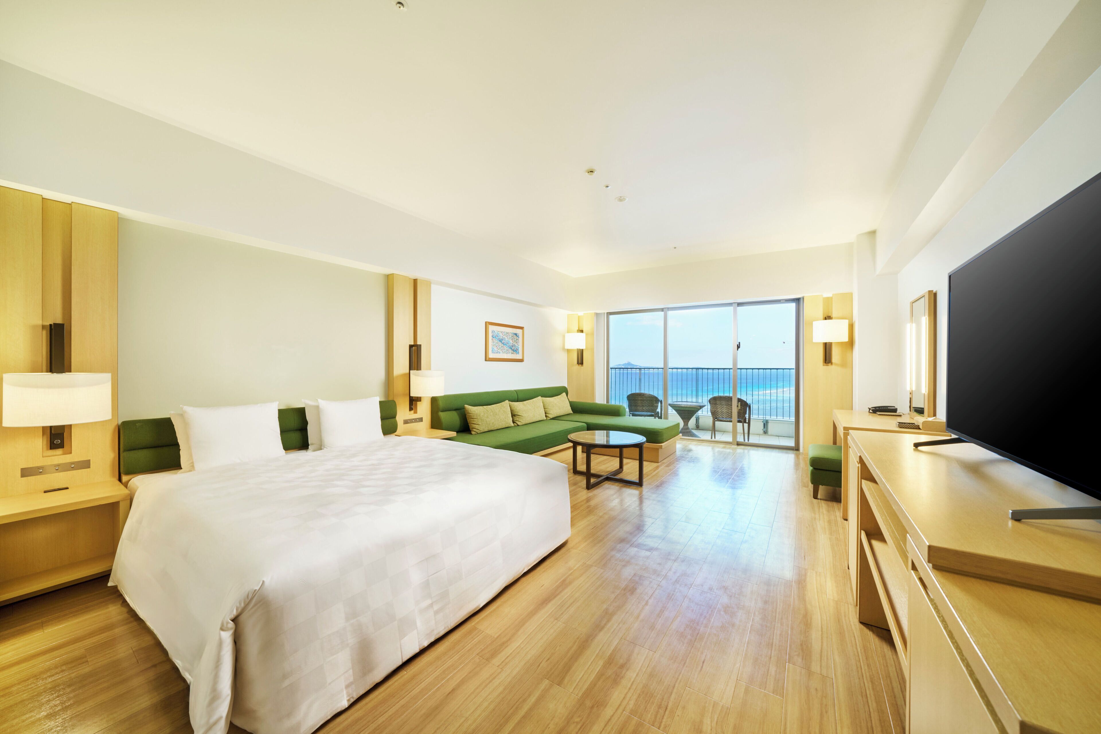 Ein Hotelzimmer mit einem großen Bett, einem grünen Sofa, einem kleinen Tisch, einem Fernseher und einem Balkon mit Blick.