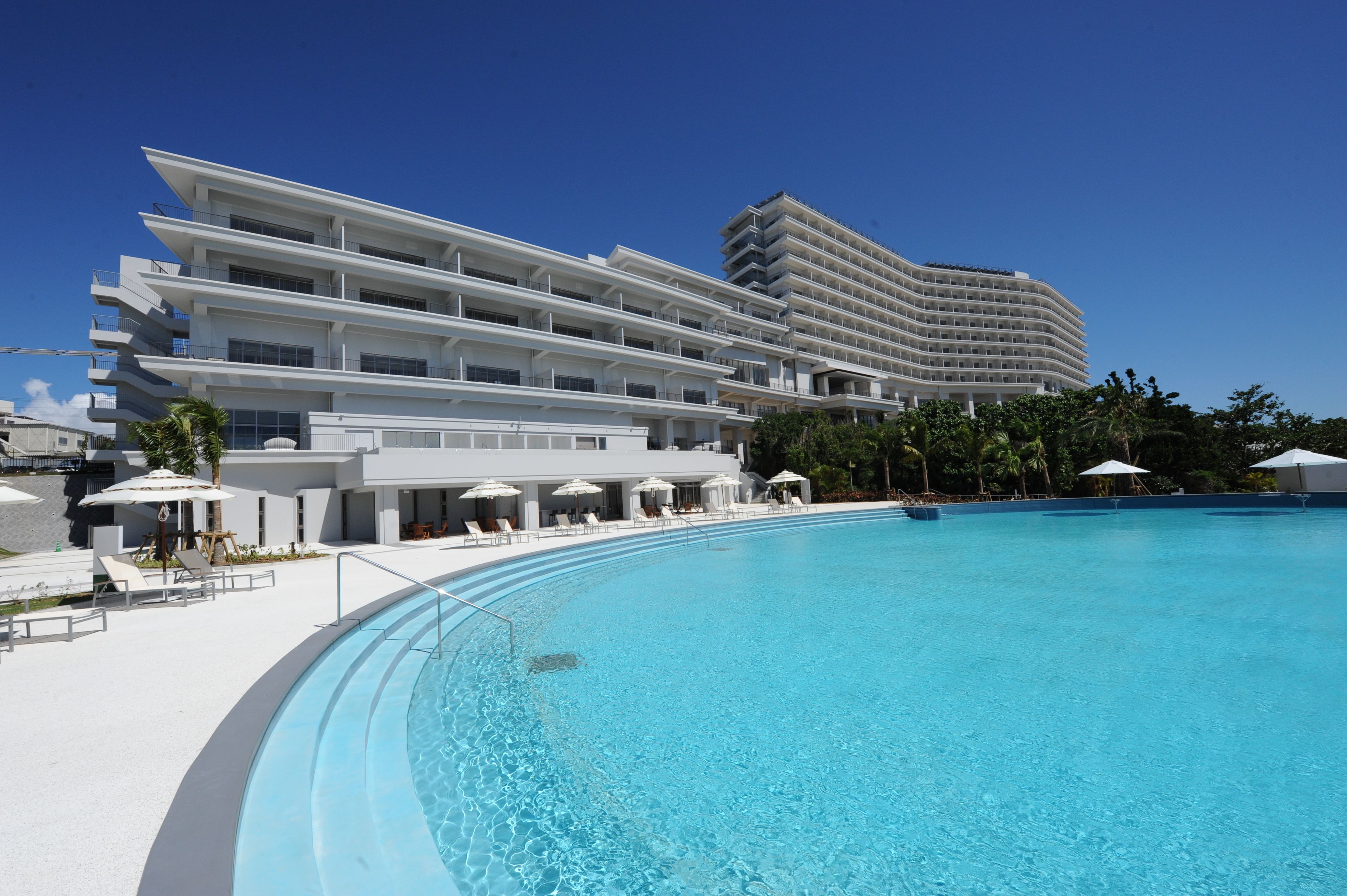 Un hotel moderno con una piscina grande, varios balcones y tumbonas.