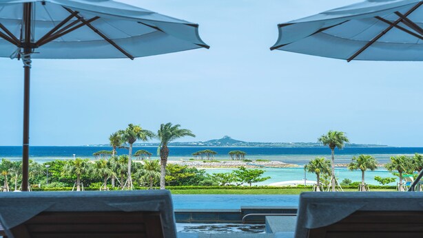 A poolside view with two umbrellas, a beach, palm trees, and a distant mountain.