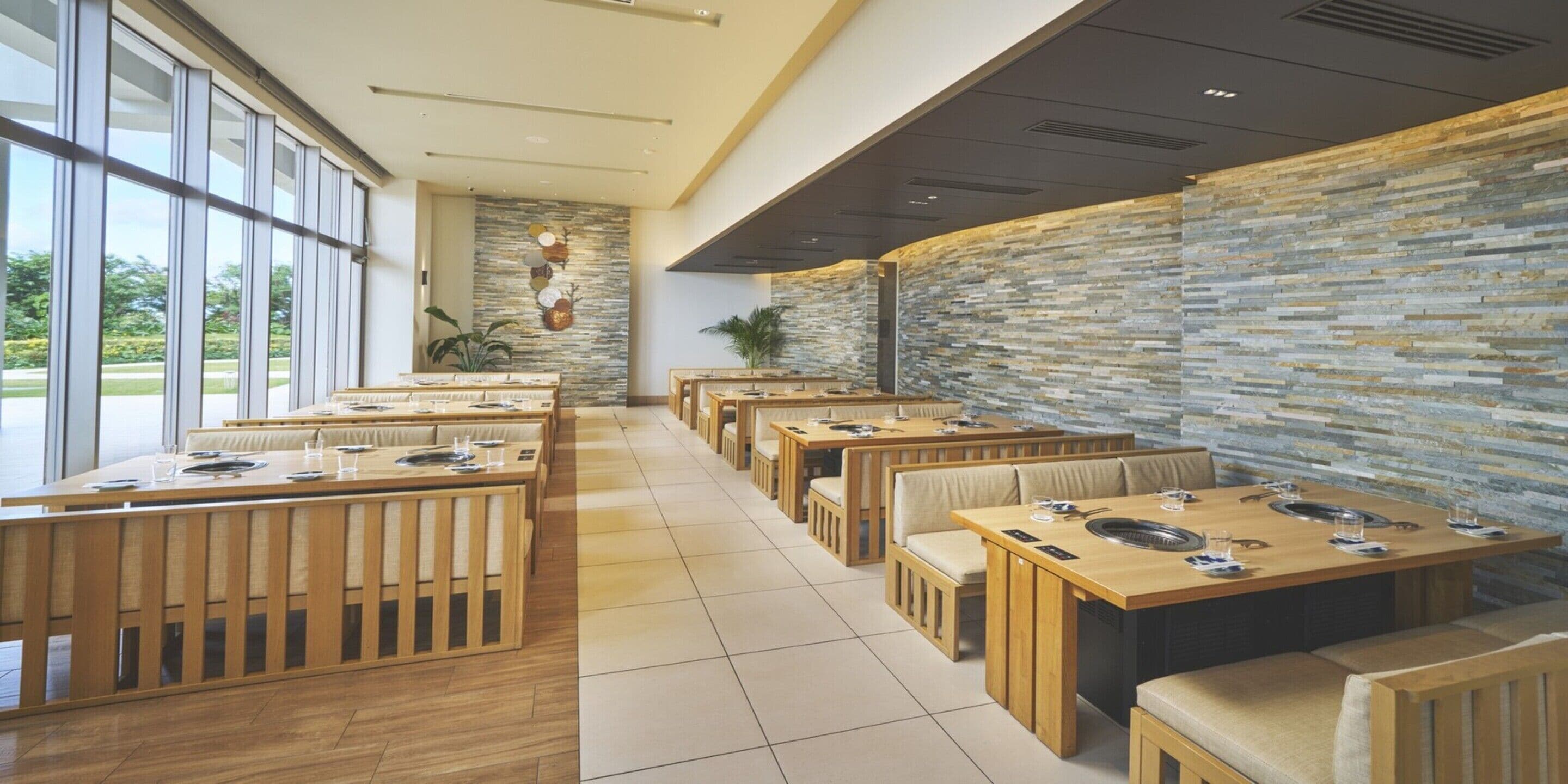 A modern restaurant with wooden tables and benches, stone walls, and large windows overlooking a garden.