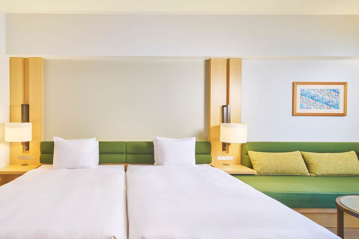Un lit double avec du linge de lit blanc, un canapé vert avec des coussins verts et deux lampes de chevet sur des tables de nuit en bois.
