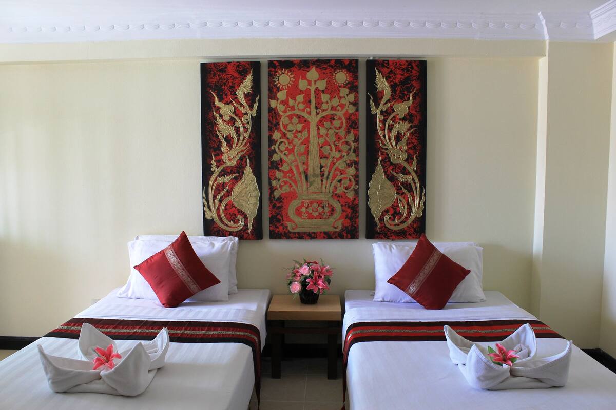 Une chambre d’hôtel avec deux lits, chacun agrémenté de draps blancs et d’oreillers rouges, une petite table sur laquelle est posé un vase de fleurs, et une décoration murale au-dessus des lits.