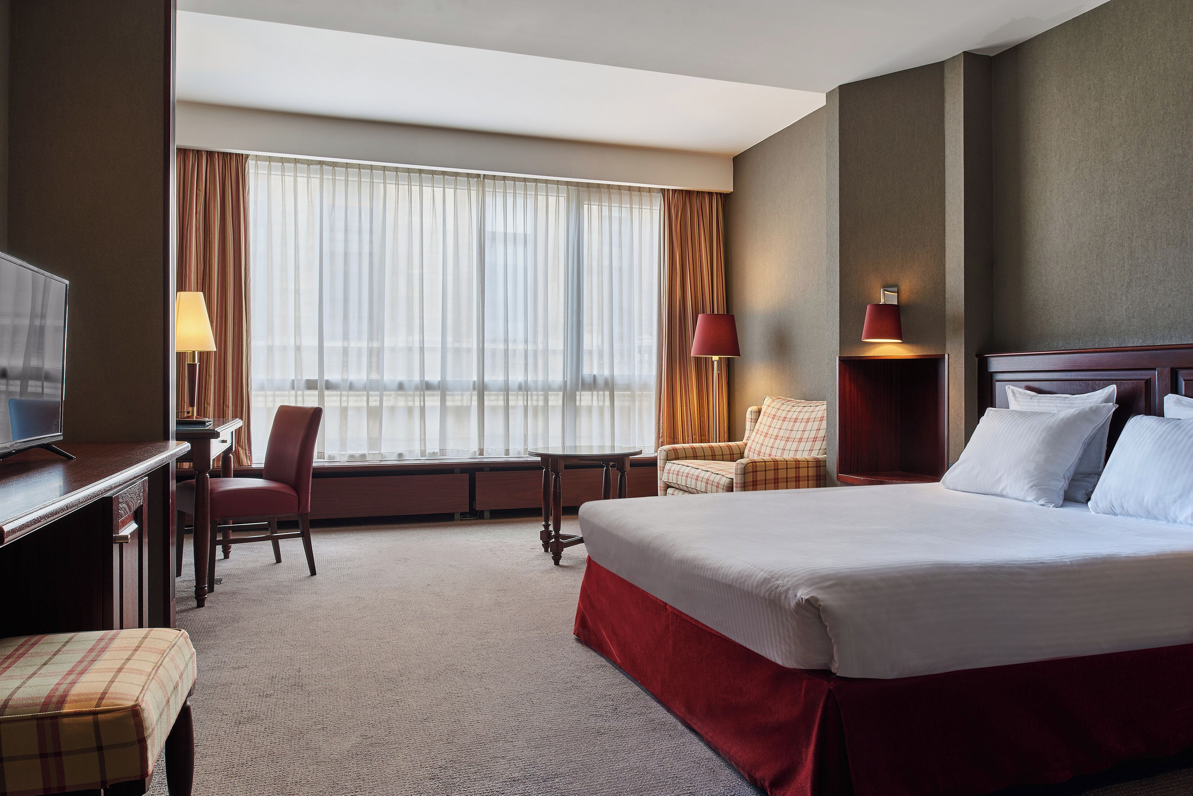 A hotel room with a large bed, a desk with a chair, a TV, and a view of the city through the window.