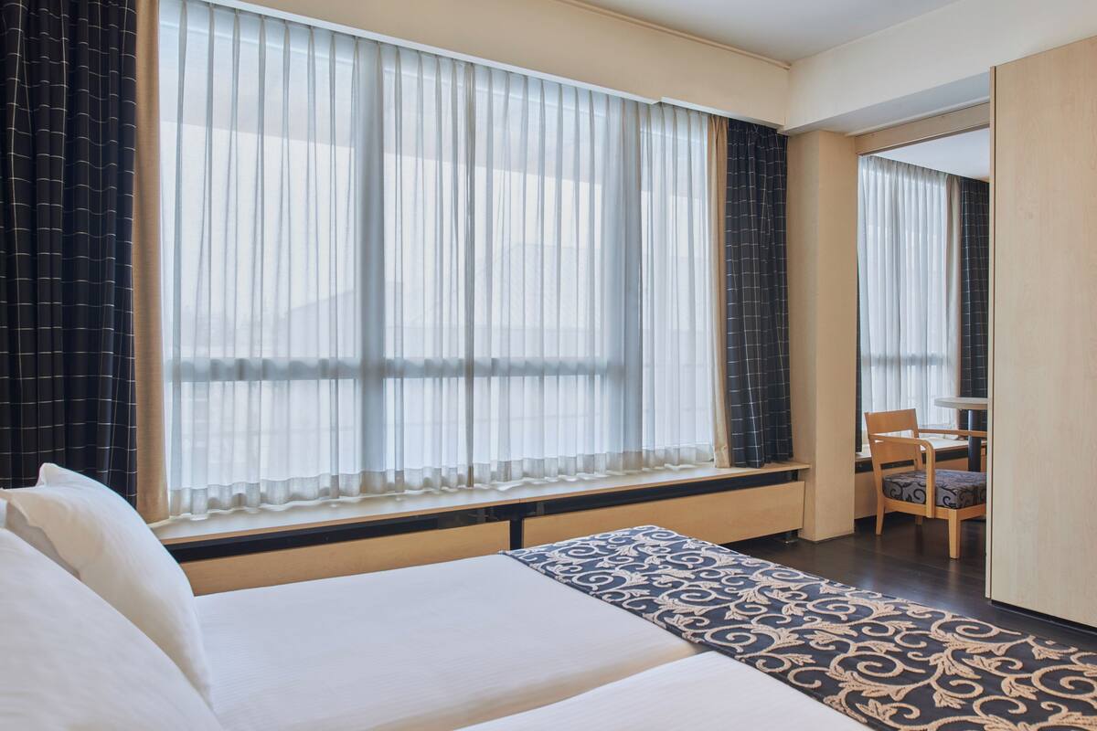 A hotel room with a large bed, a desk with a chair, and a window with sheer curtains.