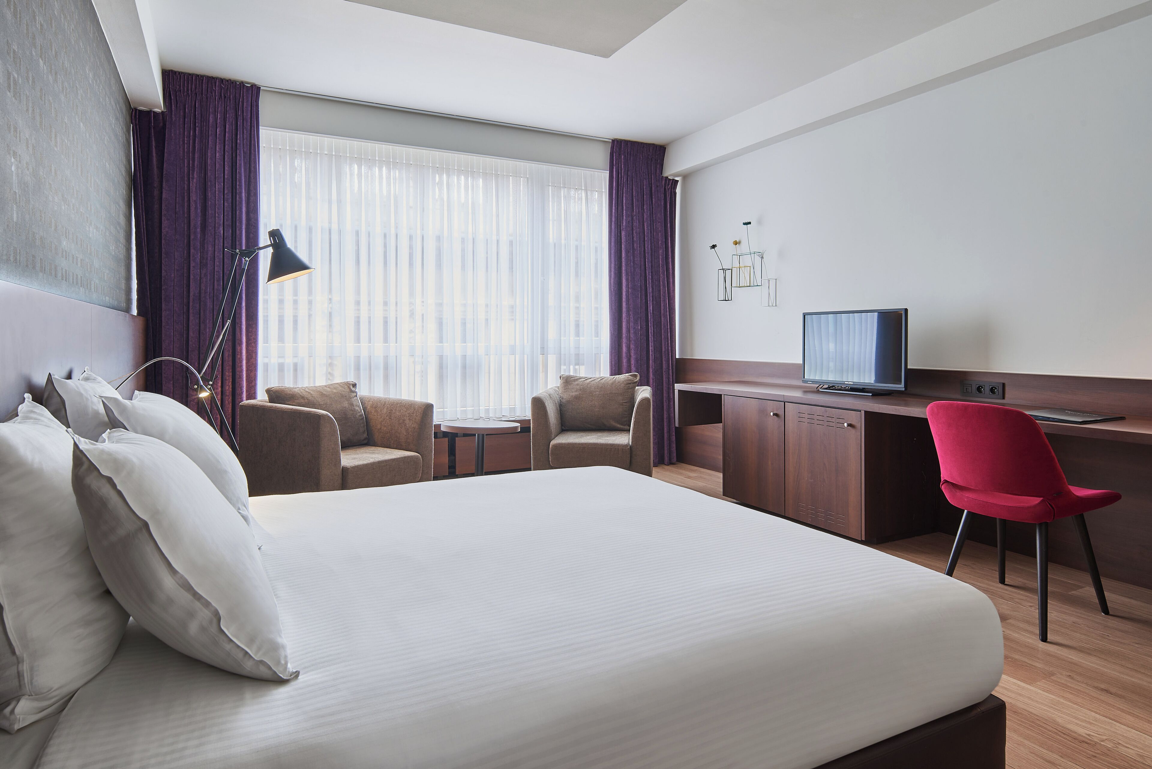 Habitación de hotel con una cama grande, dos sillones, un escritorio con televisor y una lámpara de escritorio.