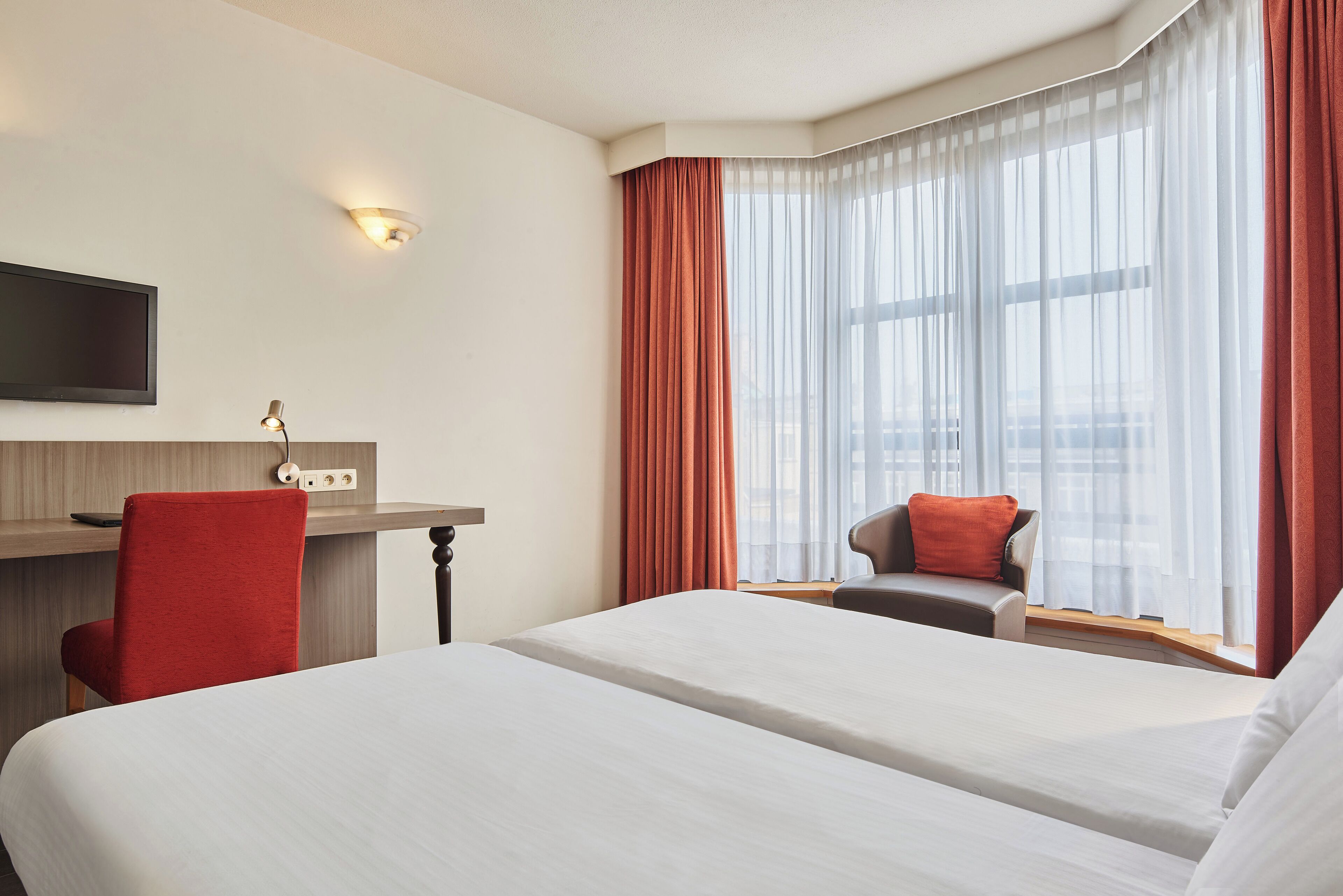 A hotel room with a bed, a desk, a chair, a TV, and a window with curtains.