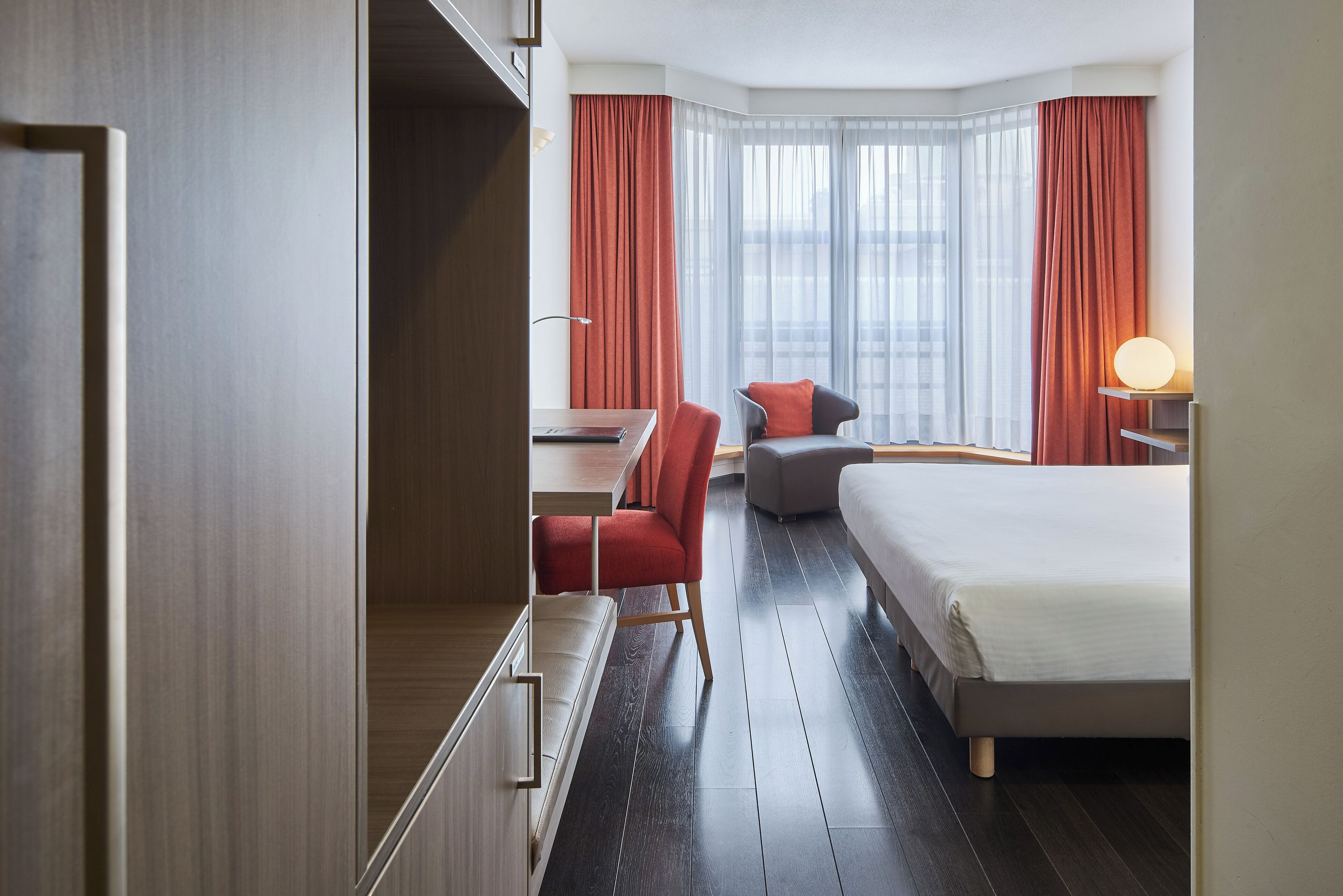 A hotel room with a bed, a desk, a chair, and a large window with curtains.