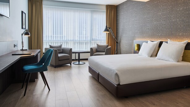 A modern hotel room with a large bed, two armchairs, a desk, and a view of the city through large windows. - Theater Hotel (Antwerp)