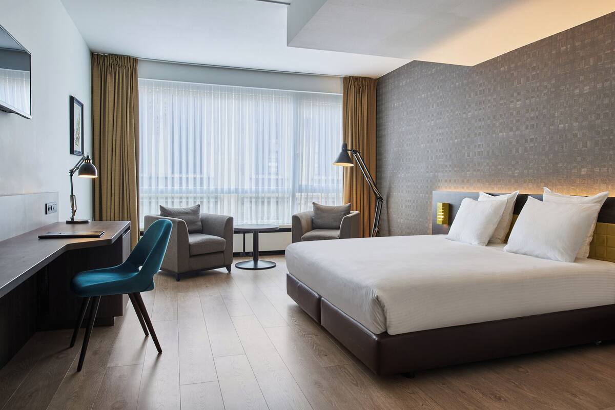 A modern hotel room with a large bed, two armchairs, a desk, and a view of the city through large windows.