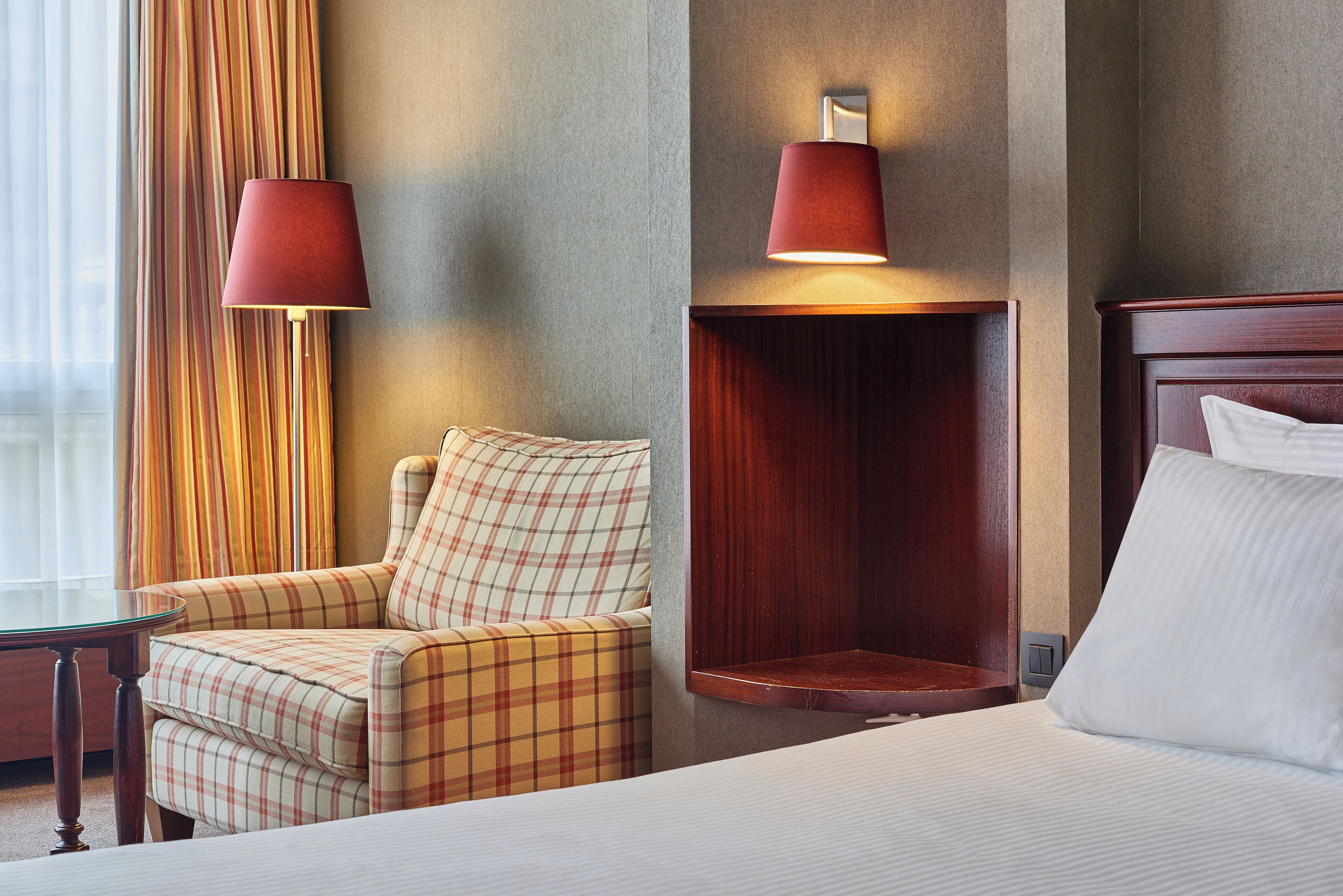 A hotel room with a bed, a plaid armchair, a small table, and two wall-mounted lamps.