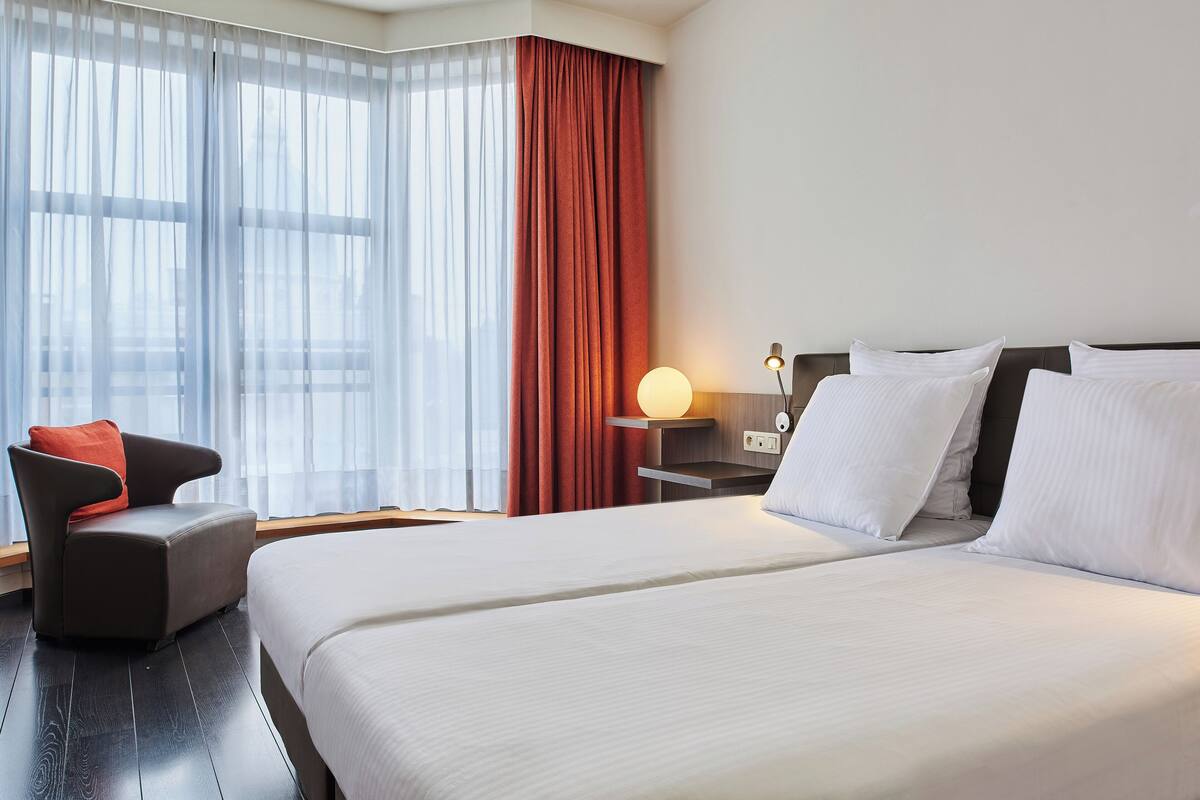 A hotel room with a large bed, a chair, a nightstand with a lamp, and a window with sheer curtains and a solid red curtain.