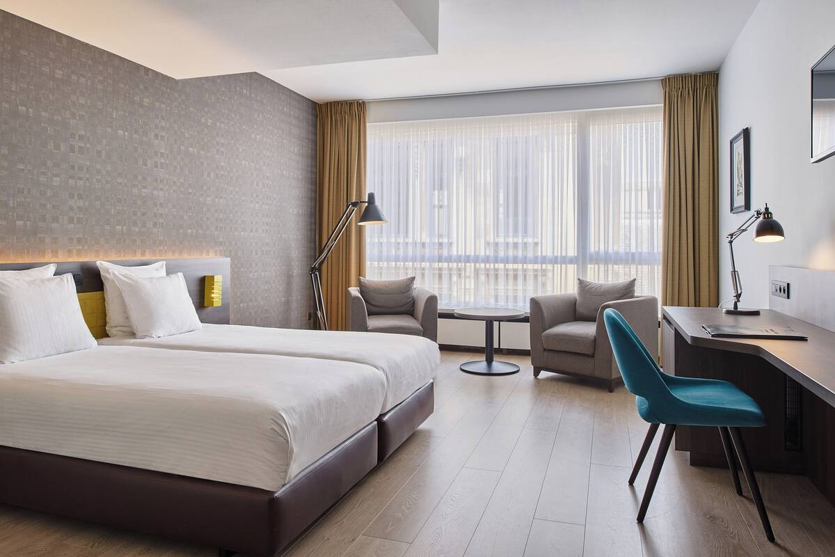 A hotel room with a large bed, a desk, two chairs, and a view of the city through large windows.
