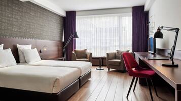 Habitación de hotel con una cama grande, dos sillones, un escritorio y un ventanal con cortinas.