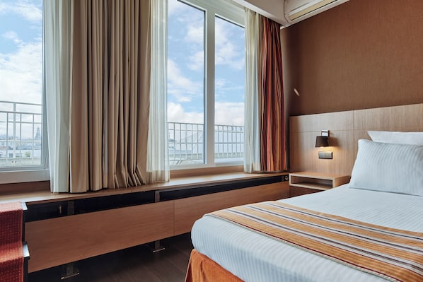 Habitación de hotel con una cama grande, una mesita de noche con lámpara, una ventana con cortinas y vistas a la ciudad.
