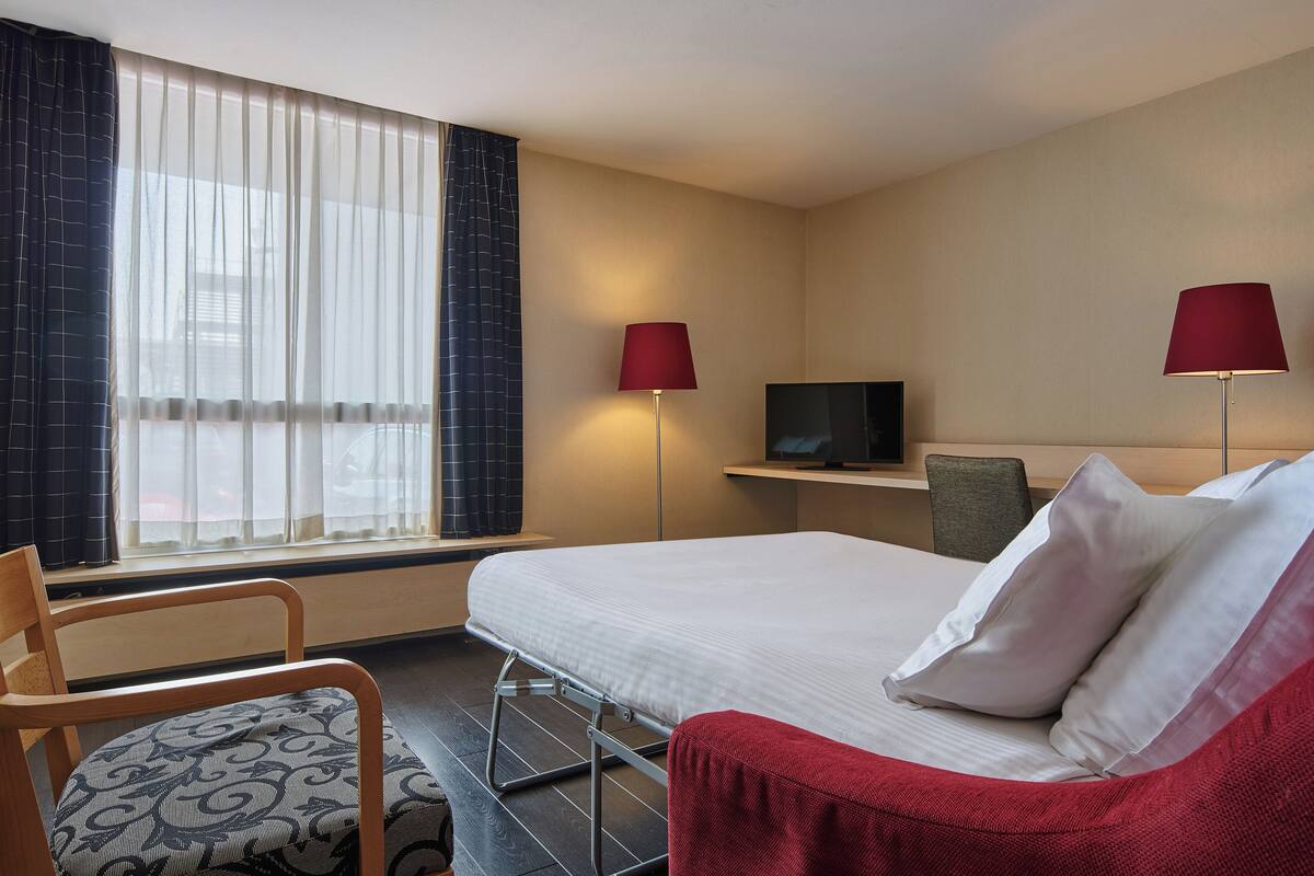 A hotel room with a bed, a chair, a TV, and a window with curtains.