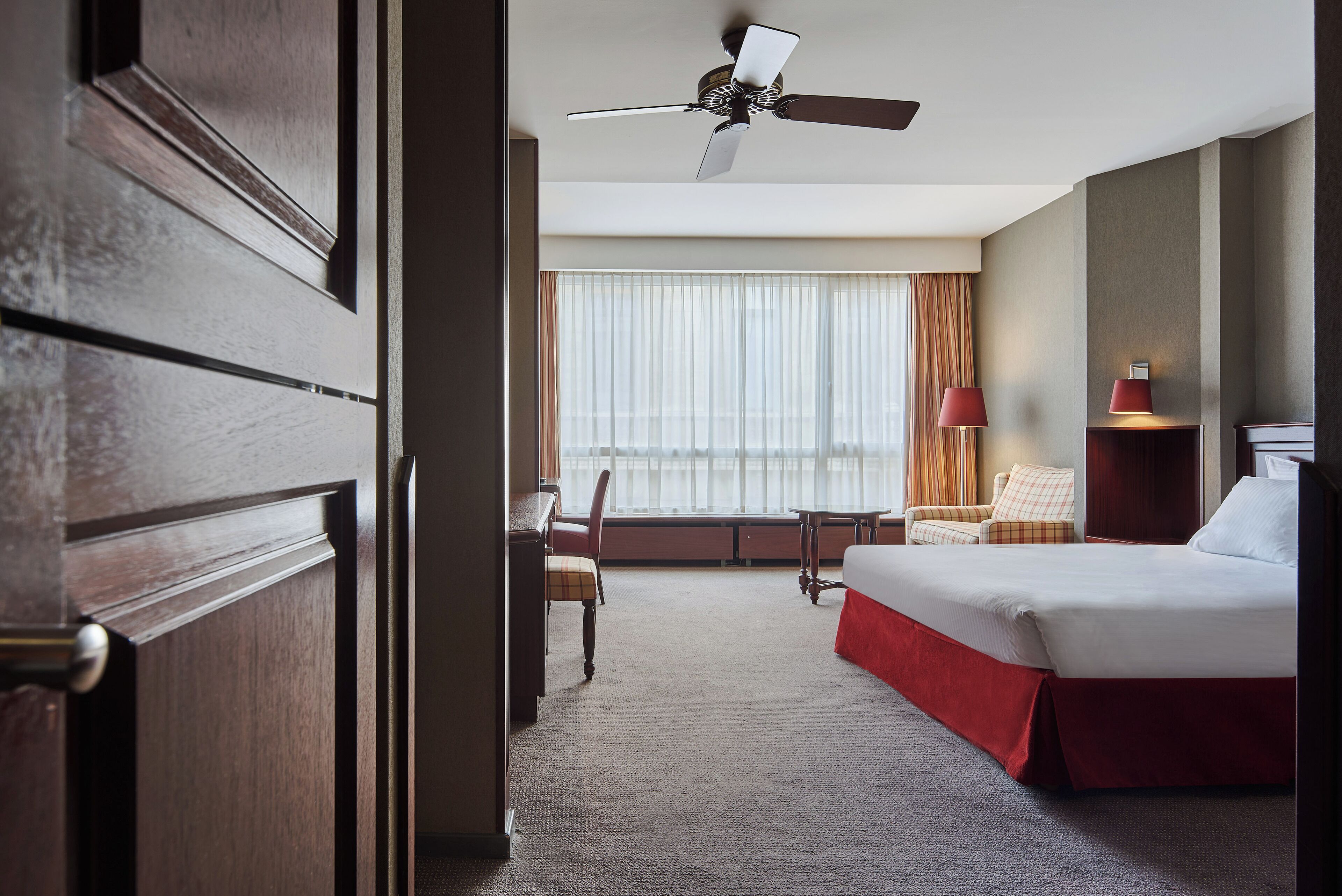 A hotel room with a bed, a desk, a chair, a window with curtains, and a ceiling fan.