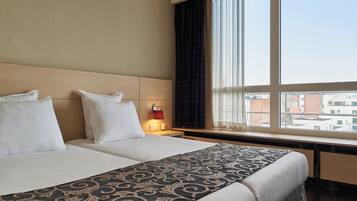 Ein Hotelzimmer mit zwei Betten, einem großen Fenster mit Vorhängen, einem Nachttisch mit Lampe und Blick auf Gebäude.