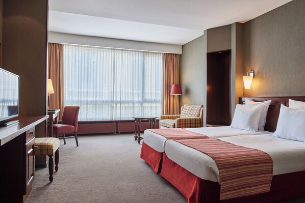 A hotel room with two beds, a desk, a chair, a TV, and a large window with curtains.