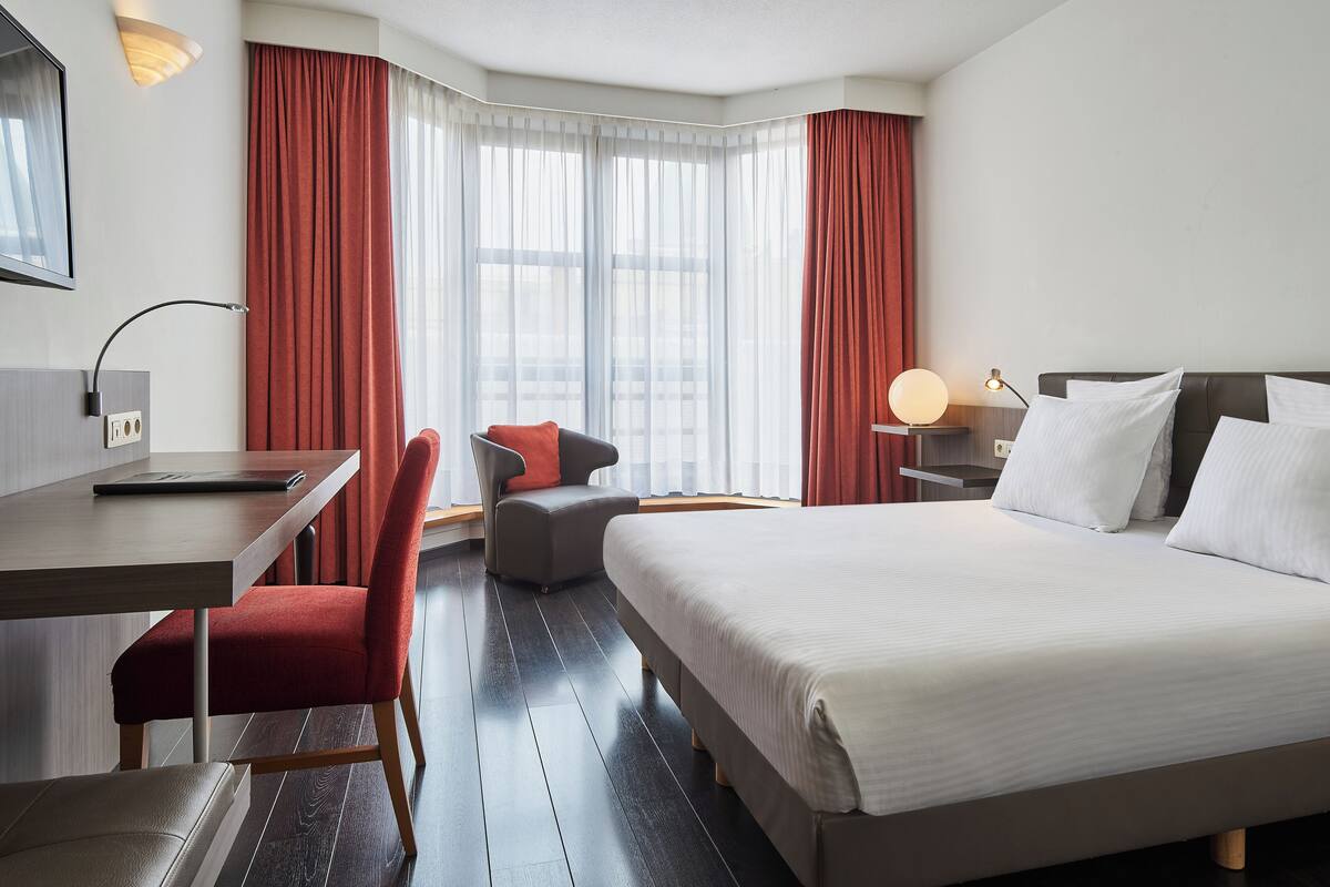 A hotel room with a bed, a desk, a chair, and a nightstand with a lamp.