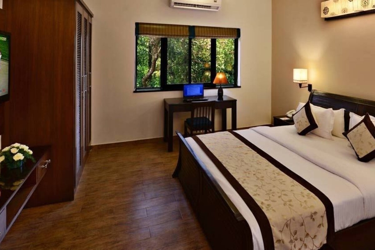 Habitación de hotel con cama, lámparas de noche, escritorio con computadora, televisor y ventana con vista a árboles.