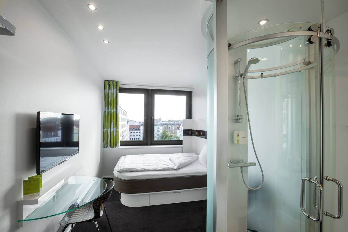 Ein modernes Hotelzimmer mit Duschkabine aus Glas, einem Bett mit Matratze und Kissen, einem Glastisch mit Stuhl, einem an der Wand befestigten Flachbildfernseher und einem Fenster mit Vorhängen.