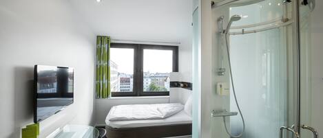Ein modernes Hotelzimmer mit Duschkabine aus Glas, einem Bett mit Matratze und Kissen, einem Glastisch mit Stuhl, einem an der Wand befestigten Flachbildfernseher und einem Fenster mit Vorhängen.