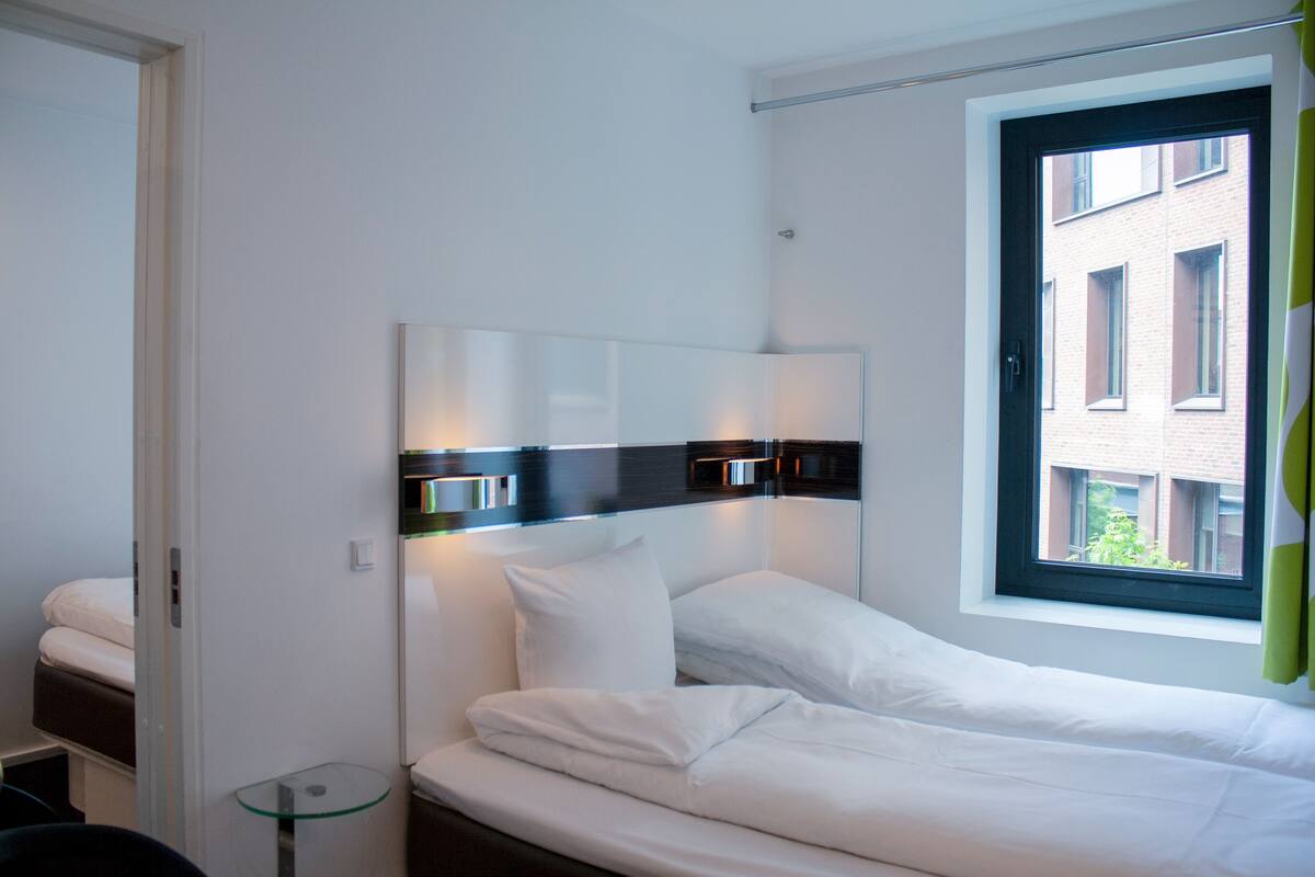 Ein modernes Hotelzimmer mit einem ordentlich bezogenen Bett, einem Nachttisch und einem Fenster mit Blick auf ein Gebäude.