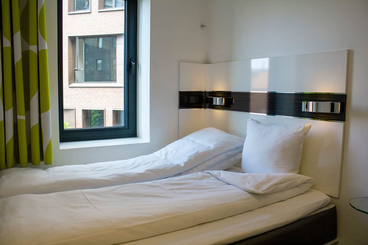 Ein Einzelbett mit weißer Bettwäsche, ein Fenster mit Vorhängen und eine Wandleuchte.