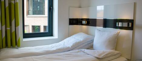 Een enkel bed met witte lakens, een raam met gordijnen en een wandlamp.