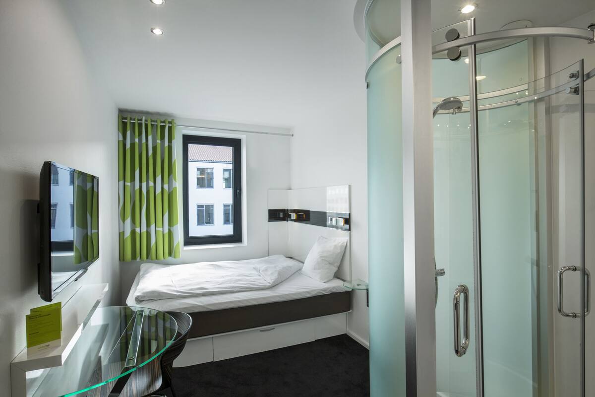 Ein modernes Hotelzimmer mit einem Bett, einem Glastisch, einem Fernseher, einer Dusche und einem Fenster mit grünen Vorhängen.
