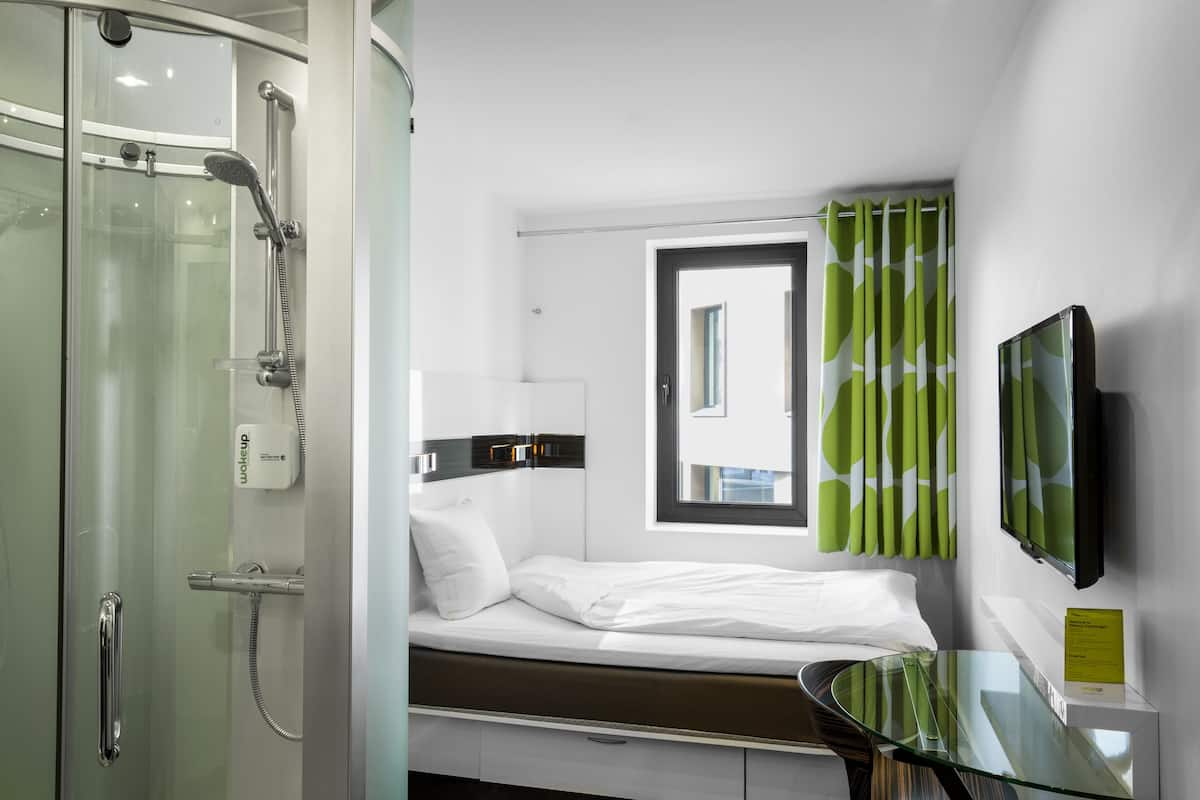Ein modernes Hotelzimmer mit Duschkabine aus Glas, einem Bett mit weißer Bettwäsche, einem Glastisch und einem an der Wand befestigten Fernseher.