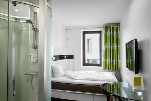 A modern hotel room with a glass shower enclosure, a bed with white linens, a glass table, and a television mounted on the wall.