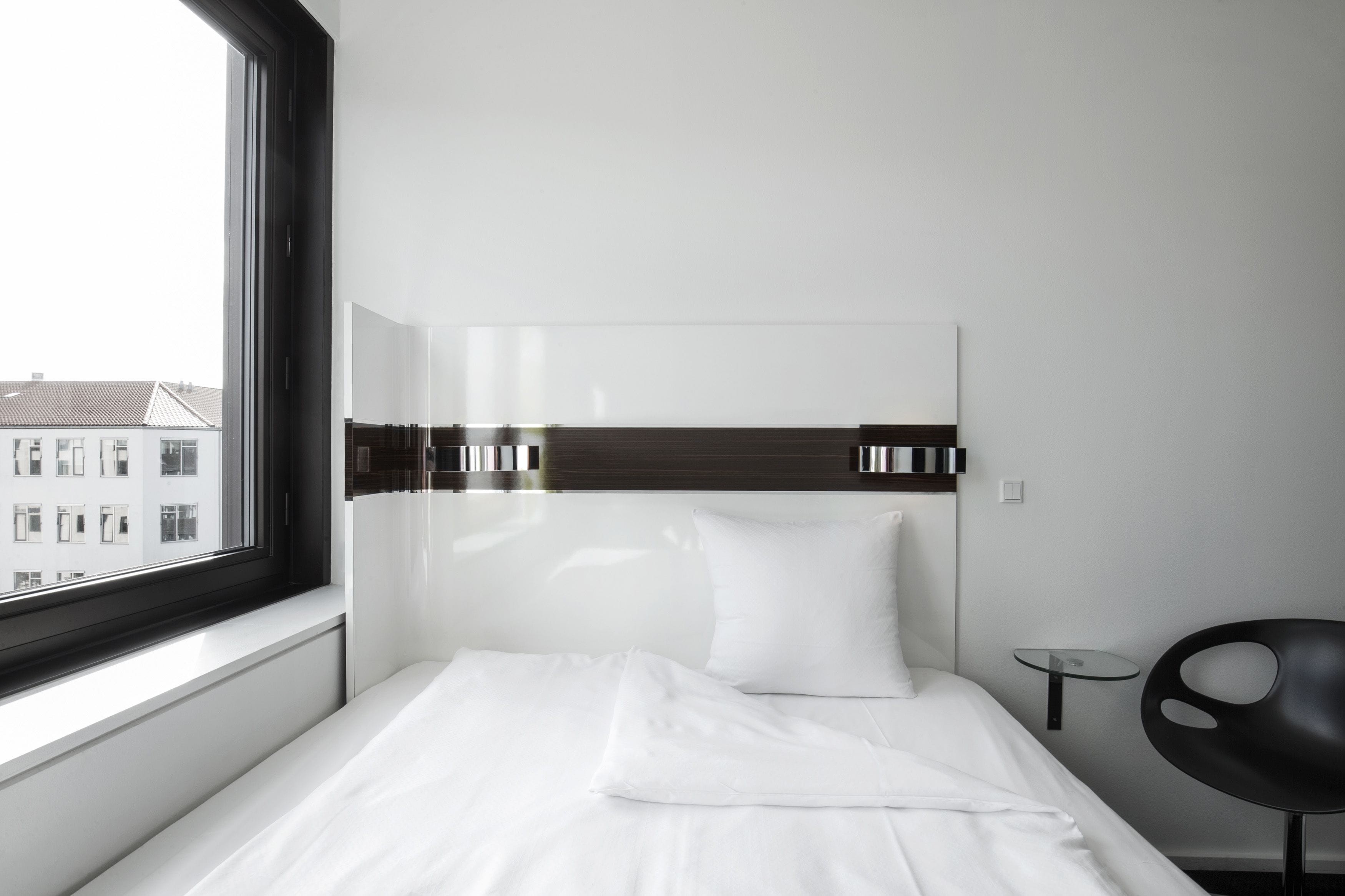 A modern hotel room with a large window, a bed with white linens, a black headboard, and a small side table.
