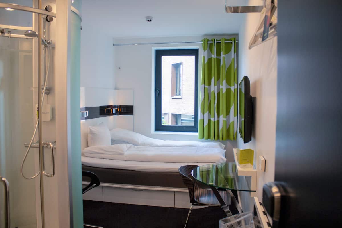 Ein Hotelzimmer mit Bett, Fenster, Dusche, Stuhl und einem an der Wand befestigten Fernseher.
