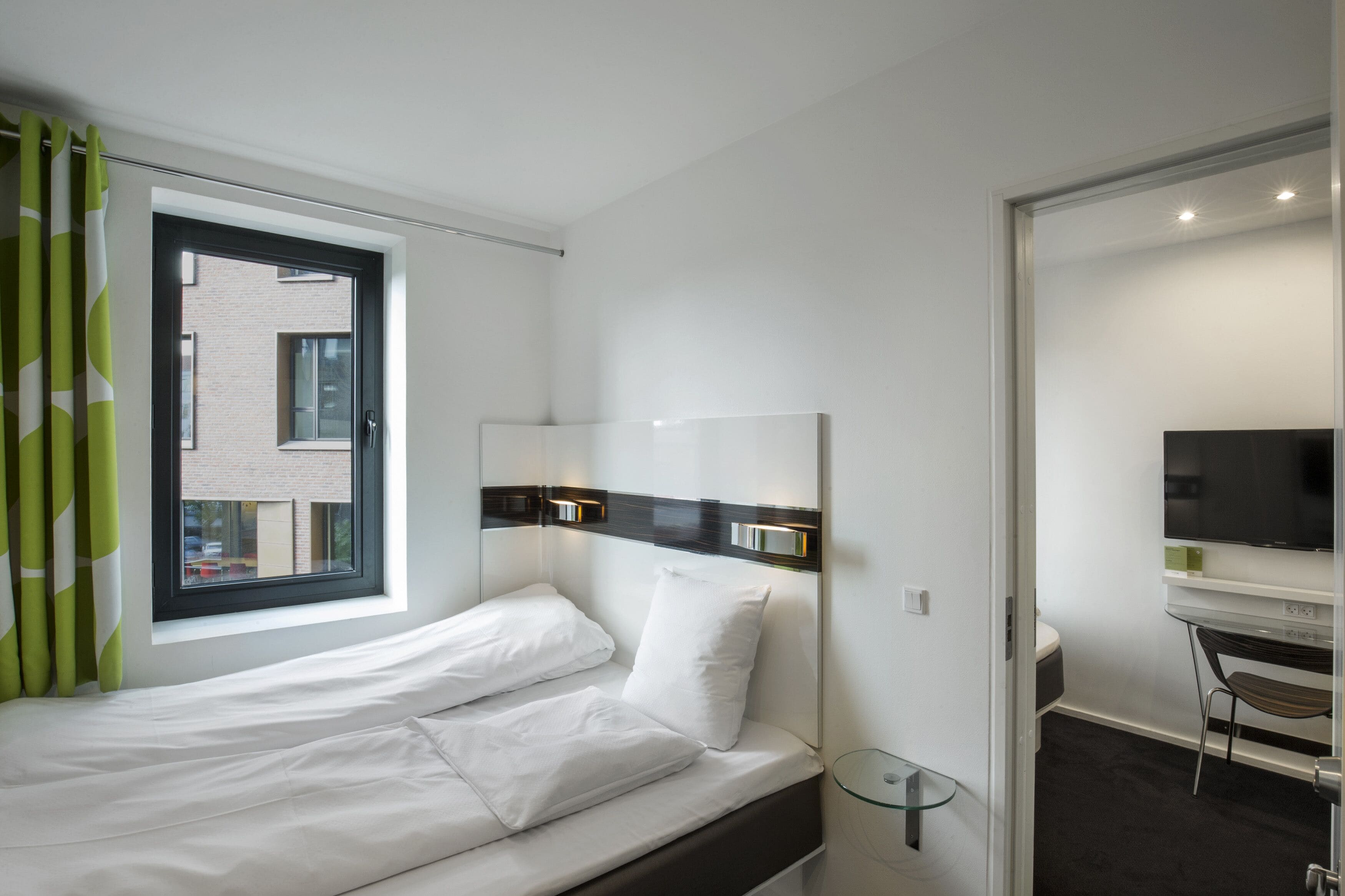 A modern hotel room with a bed, a desk, a TV, and a window with curtains.