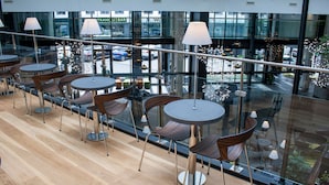 A modern indoor seating area with round tables and chairs, large windows offering views of the street, and a glass railing overlooking the seating area.