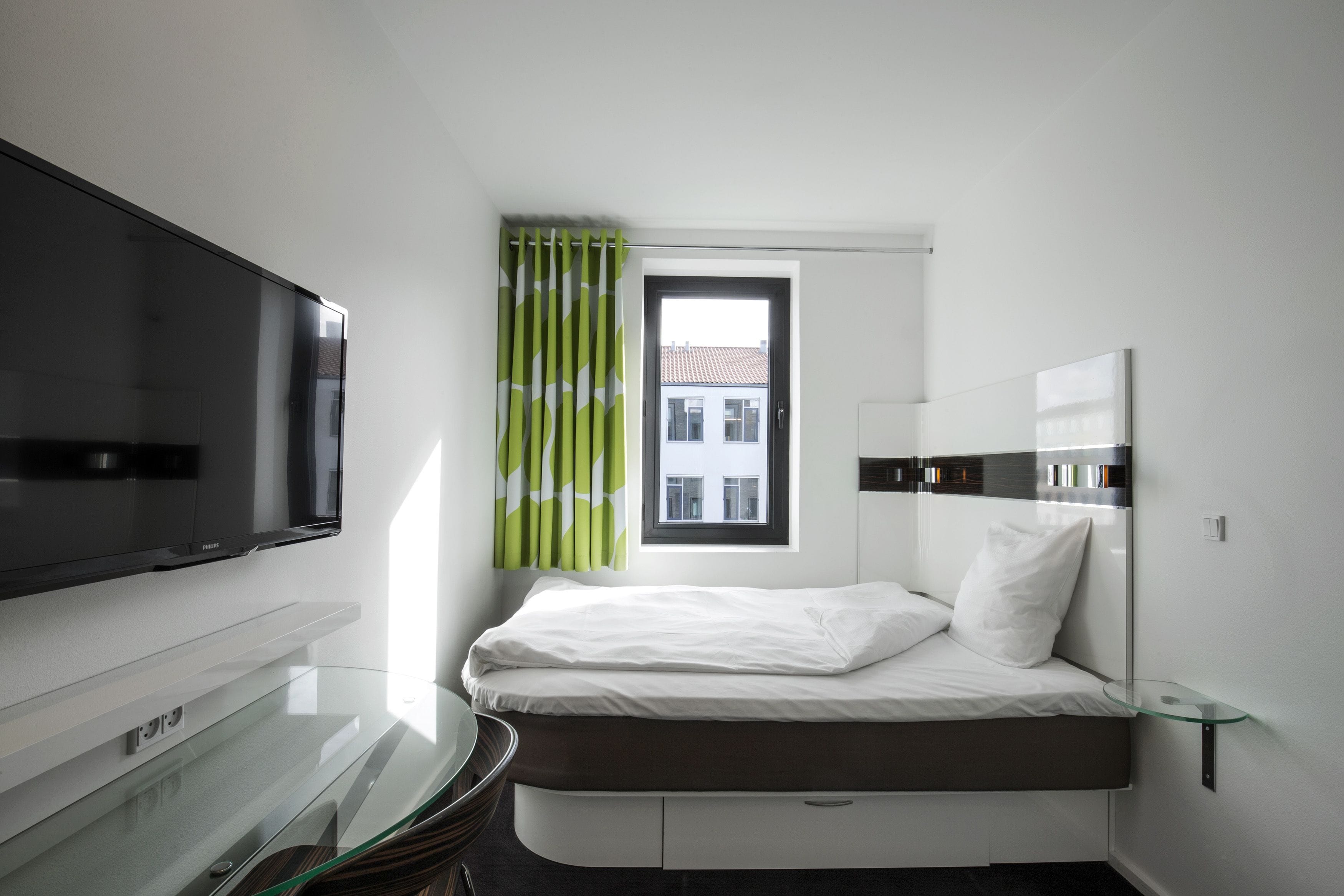 A modern hotel room with a flat-screen TV mounted on the wall, a bed with white bedding, a glass coffee table, and a window with green curtains.
