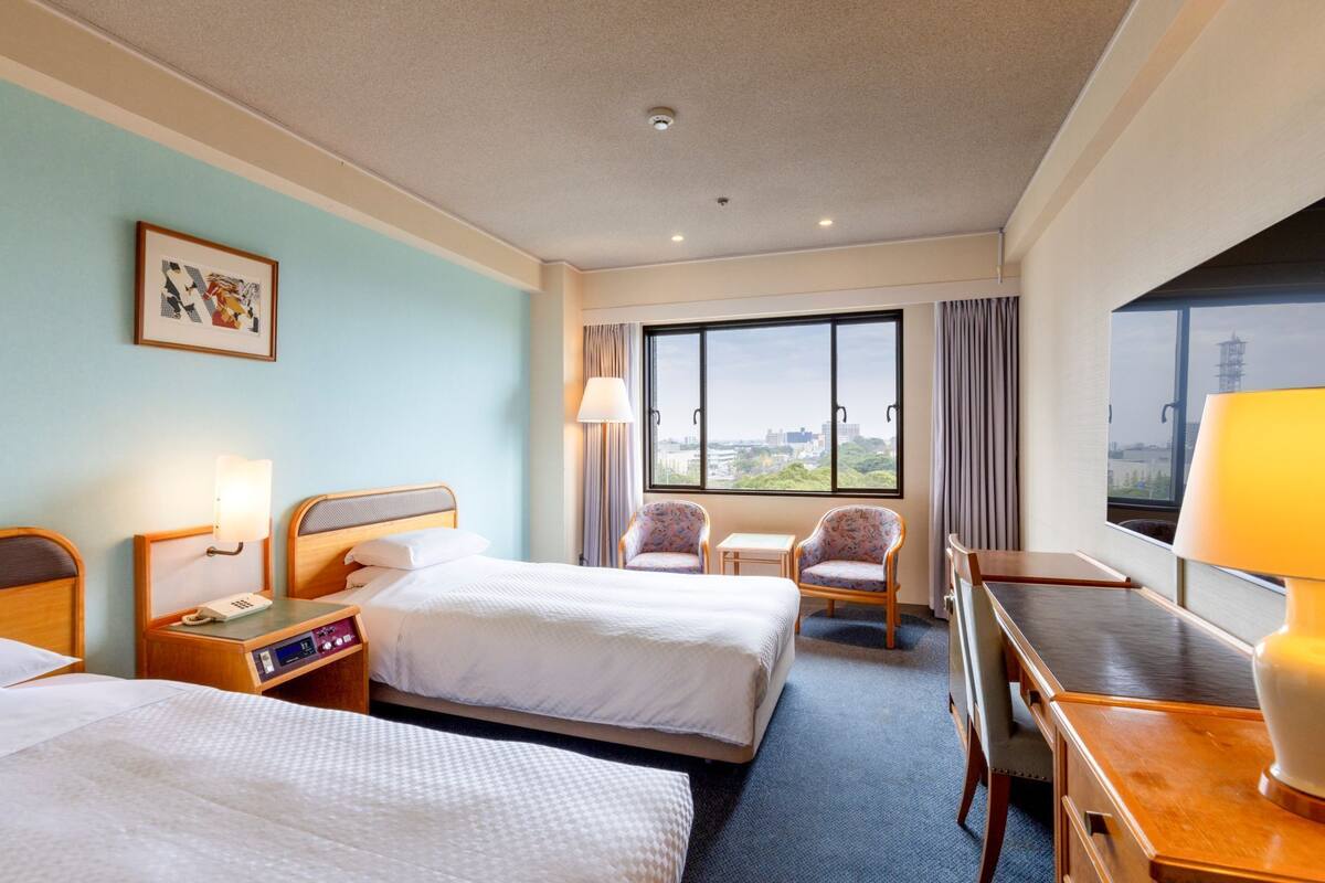 Une chambre d’hôtel avec deux lits, un bureau, des chaises, une fenêtre donnant sur la ville et un tableau au mur.