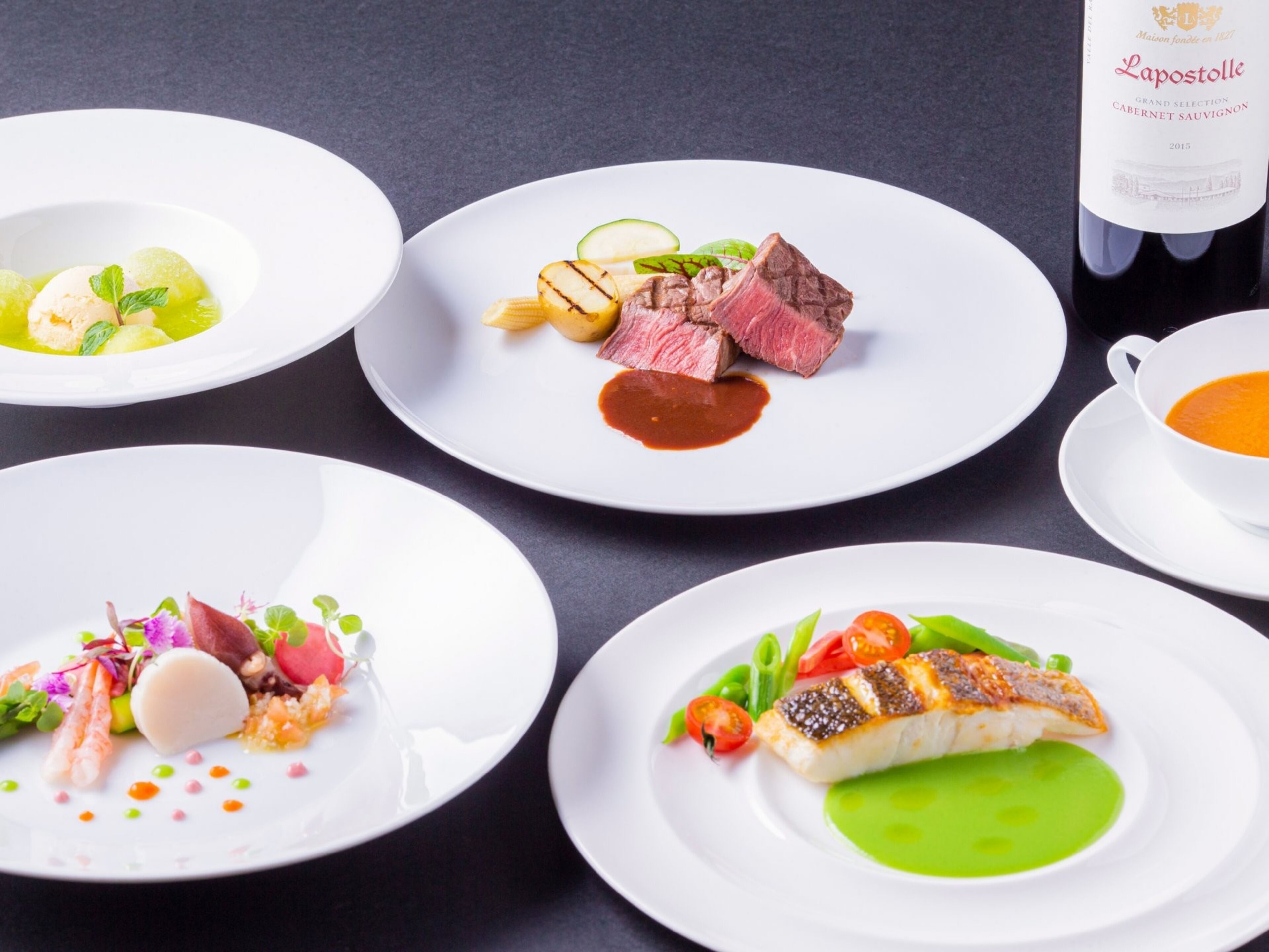 A table with four plates of food, a bottle of wine, and a cup of soup.